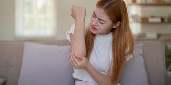 Young woman feeling elbow pain while sitting on sofa at home.