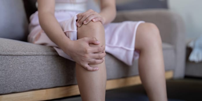 Young woman feeling knee pain while sitting on sofa at home.