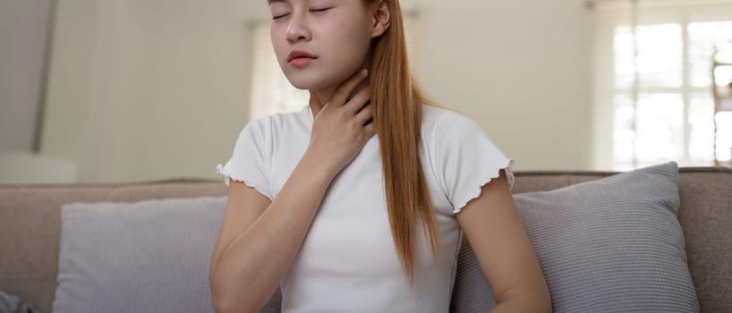 Young woman experiencing throat pain while sitting on a sofa at home.
