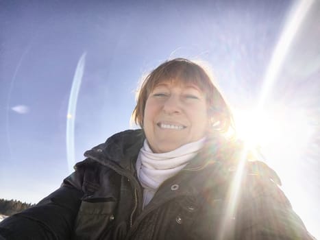 A cheerful middle aged woman in a winter coat taking selfie on nature outdoors in sunny day with blue sky