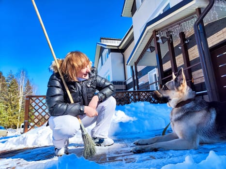 A girl or a woman with a snow broom at home and a large German Shepherd dog. Woman Clearing Snow at Home With Her German Shepherd