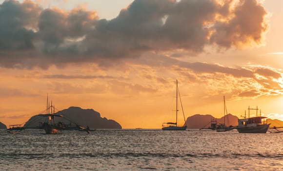 Multiple boats are anchored in a calm bay under a vibrant sunset. The sky glows with shades of orange and gold, while dark clouds create dramatic contrasts.