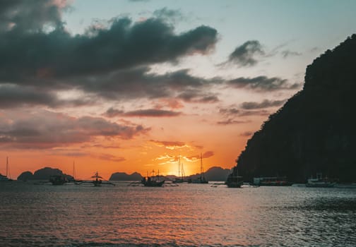 Multiple boats are anchored in a calm bay under a vibrant sunset. The sky glows with shades of orange and gold, while dark clouds create dramatic contrasts.