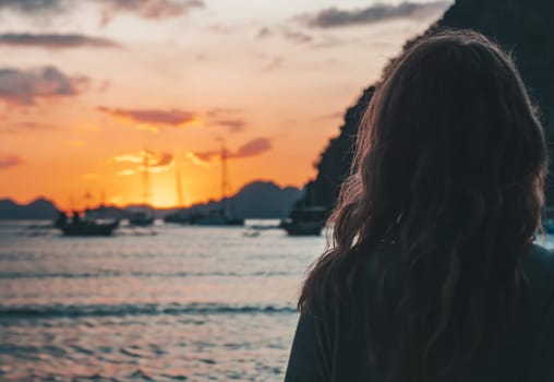 A woman stands on the beach in El Nido, Palawan, watching a stunning sunset over the calm ocean waters, with boats in the distance.