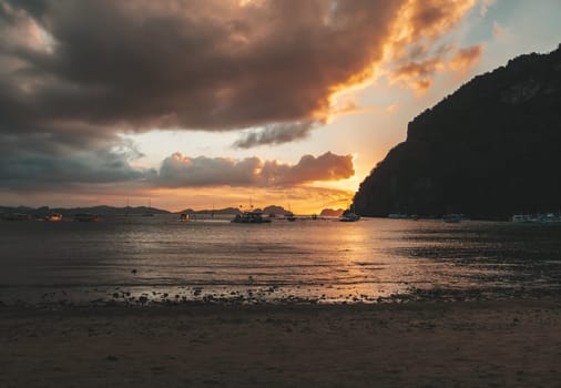 Multiple boats are anchored in a calm bay under a vibrant sunset. The sky glows with shades of orange and gold, while dark clouds create dramatic contrasts.