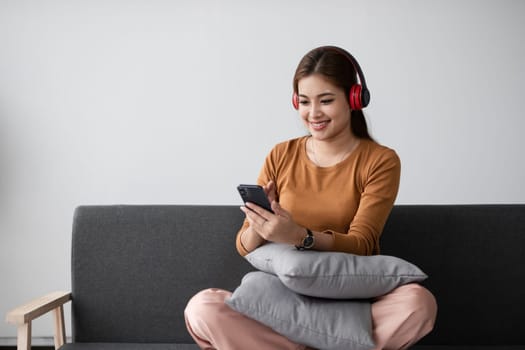 Young Asian woman spends her free time at home sitting on the sofa wearing headphones listening to music to relax and have fun..