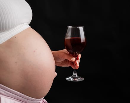 Close-up of the belly of a pregnant woman holding a glass of red wine while sitting on the sofa. Copy space