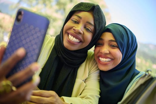 Two Middle Eastern Muslim women, adorned in hijabs, capture a moment of friendship and joy as they take selfies on a smartphone while seated in a natural setting.