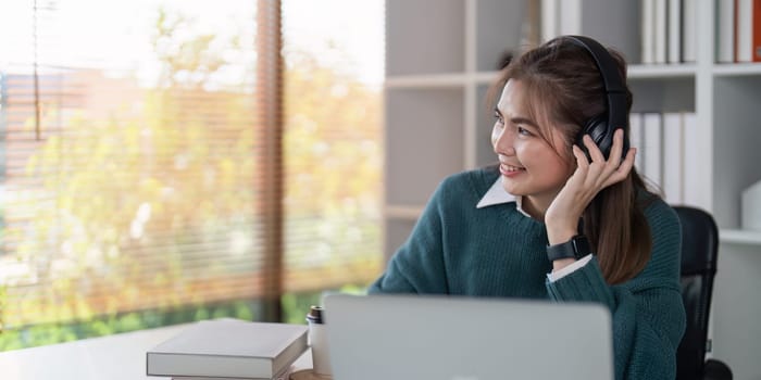 Young woman smiling while wearing headphones and using a laptop in a bright home office.