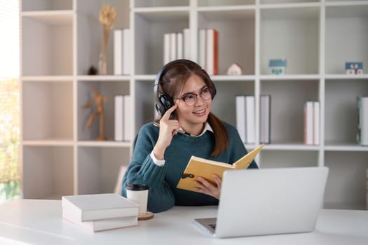 Young woman wearing headphones while studying with a book and laptop at a home office desk.