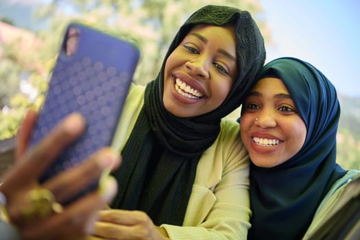 Two Middle Eastern Muslim women, adorned in hijabs, capture a moment of friendship and joy as they take selfies on a smartphone while seated in a natural setting.