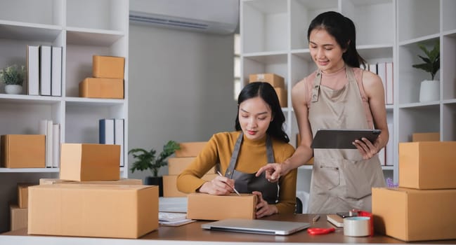 Two Asian women packing boxes and using a tablet in a home office. Concept of small business and e-commerce..