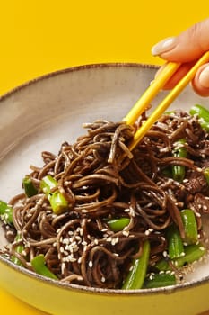 Female hand skillfully picking up delicious bite of soba noodles with beef strips and fresh green beans using chopsticks from ceramic bowl, against sunny yellow backdrop. Asian style comfort food