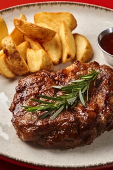 Delicious roasted pork steak with side dish of crispy country style potatoes and barbecue sauce on ceramic plate garnished with fresh aromatic rosemary against red background