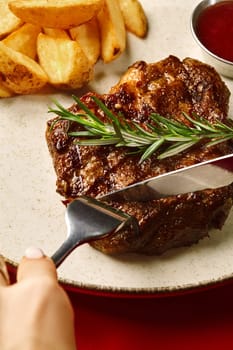 Person slicing succulent grilled pork steak topped with fresh rosemary, served on speckled plate with crispy potato wedges and barbecue sauce, cropped shot against red background