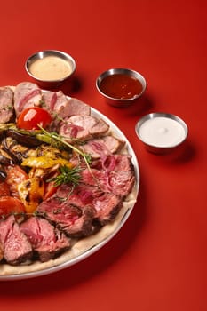 Assorted meat platter with roast beef and baked pork loin garnished with fresh aromatic herbs, served with mix of grilled vegetables and marinated onions, accompanied by dips against red backdrop