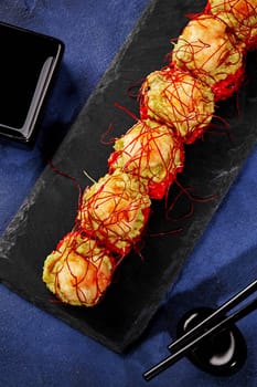 Red tobiko sushi rolls topped with avocado spread and shrimp tempura garnished with spicy togarashi threads traditionally served with soy sauce on black slate board on blue backdrop. Japanese cuisine