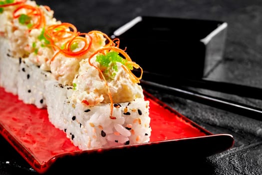 Closeup of appetizing sesame coated roll with creamy seafood topping and green tobiko roe, garnished with spicy chili shavings served on red plate with soy sauce and chopsticks on black surface