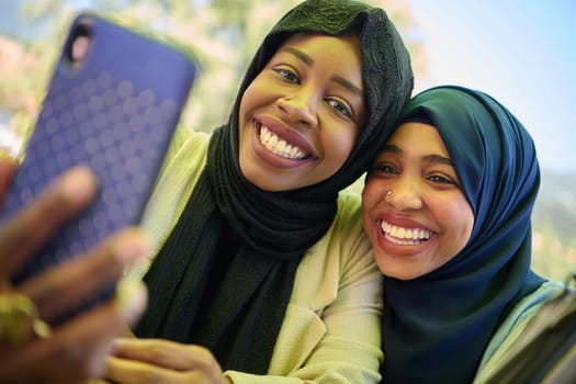 Two Middle Eastern Muslim women, adorned in hijabs, capture a moment of friendship and joy as they take selfies on a smartphone while seated in a natural setting.