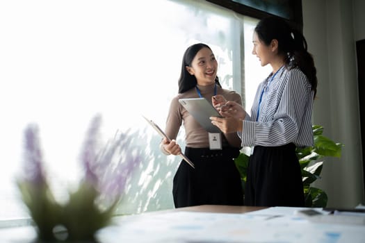 Two Asian businesswomen discussing work using a tablet in a modern office. Concept of teamwork and professional collaboration.