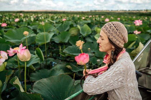 A woman is sitting in a boat in a field of pink lotus flowers. She is wearing a hat and a scarf