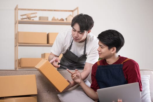 A gay couple running a small business takes order on a laptop and sells product online together in a room full of boxes..