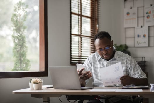 African American businessman working on paperwork, searching for information with laptop at home office.