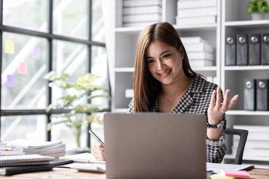 Young Asian business woman reviews company work documents, checking information on a laptop on a work desk in a white office..