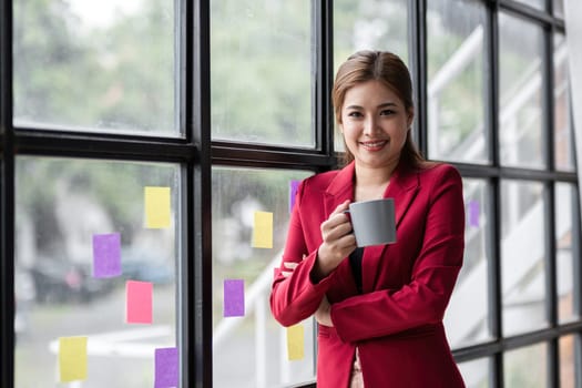 Confident smiling business woman stands holding a coffee mug and looks out the window..