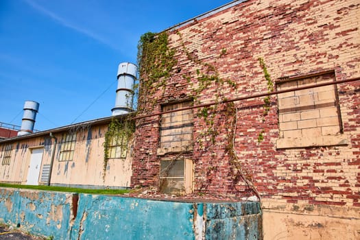 Sunlit abandoned industrial building in Warsaw, Indiana, with ivy-clad brick walls and rusted vents.