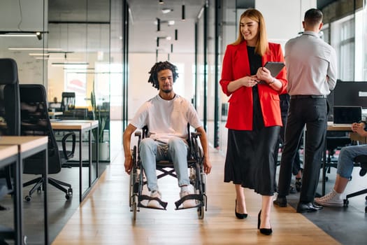 A business leader with her colleague, an African-American businessman who is a disabled person, pass by their colleagues who work in modern offices.