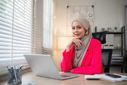 Young Muslim businesswoman wearing hijab sits working with laptop managing personal business in private office..