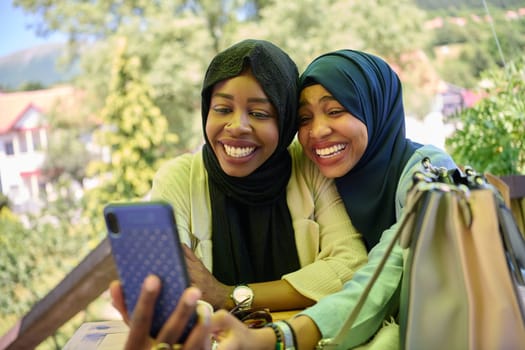 Two Middle Eastern Muslim women, adorned in hijabs, capture a moment of friendship and joy as they take selfies on a smartphone while seated in a natural setting.