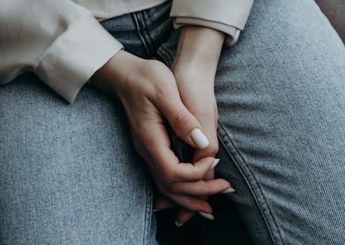 Beautiful view of the hands of a young Caucasian girl with a beautiful white manicure folded on her legs in blue jeans, spring day, top side view close-up.
