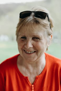 Portrait of one beautiful elderly Caucasian woman in a red sweater, sunglasses on her head with a happy smile on a spring day in a public park, close-up side view.