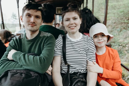 Portrait of one beautiful Caucasian family consisting of a young mother, an adult son and a little niece, looking with a smile at the camera and sitting in an open carriage of a tourist train traveling through a nature reserve on a spring day in Belgium, close-up side view.