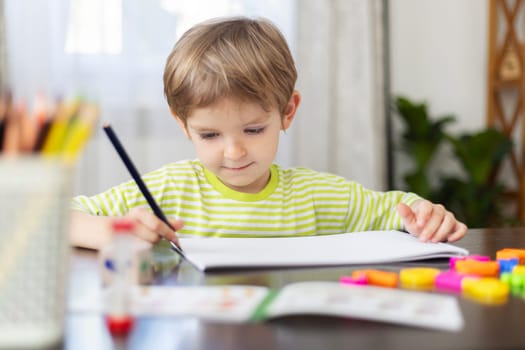 A young boy in a striped green shirt diligently paints on a white sheet of paper, surrounded by colorful crayons and a bright setting that enhances the feeling of a creative, playful childhood