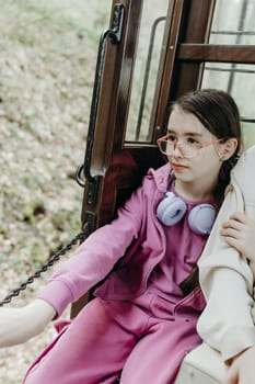 A portrait of one beautiful Caucasian girl in glasses, a light lilac tracksuit, sits on the edge of an open wooden retro carriage of a tourist train traveling through a nature reserve and looks to the side, looking at the beauty of nature on a spring day in Belgium, close-up side view.