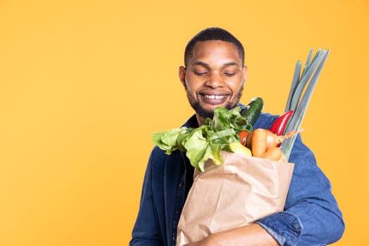 Positive enthusiastic guy smiling at his ethically sourced groceries, feeling pleased with his farmers market shopping session. Vegan person carrying his paper bag full of natural organic produce.