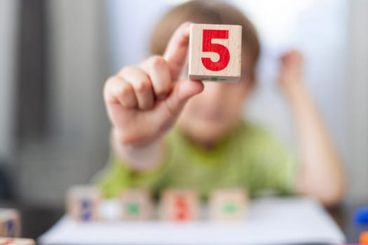 A focused young child in a striped shirt holds up a wooden block with the number 5, blurred colorful number blocks in the background, emphasizing early education in mathematics