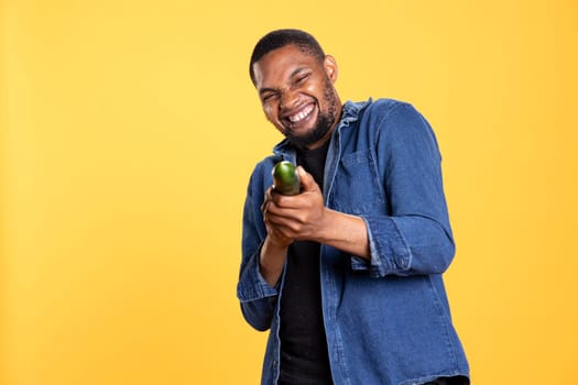 African american funny man pretending to use a cucumber as a shotgun in studio, playing around and having fun with eco friendly vegetables. Model acting goofy, zero waste concept.