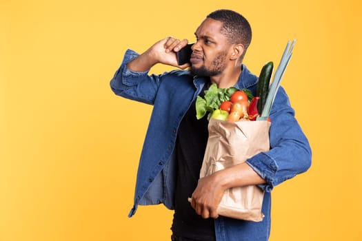 Angry displeased person starting an argument on phone call in studio, feeling furious after hearing something bad. Man with bag of groceries fighting with a friend on smartphone line.