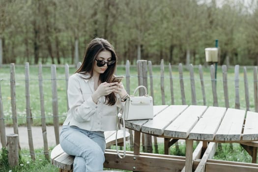 Portrait of one beautiful Caucasian brunette girl in stylish clothes with sunglasses sitting at a wooden table and browsing social networks on a smartphone in a nature reserve on a spring day in Belgium, close-up side view.