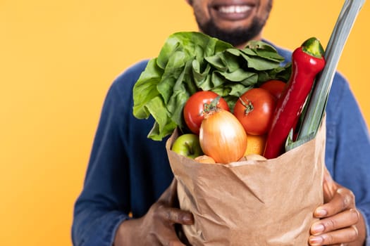 Man stands confidently with a paper bag full of fresh organic produce, grinning and presenting eco friendly vegan grocery goods. Zero waste sustainable lifestyle enthusiast. Close up.