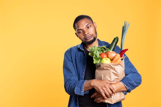 Pensive displeased young guy holds a paper bag full of bio food, shopping for eco friendly fruits and vegetables. Disappointed sad african american person buys organic produce.