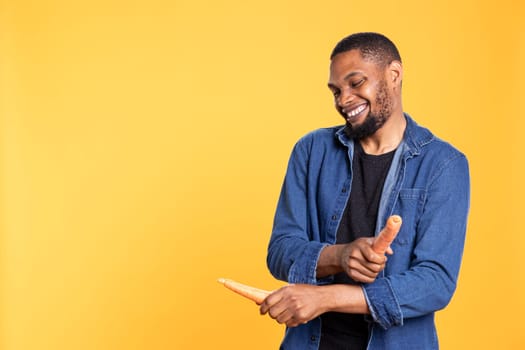 African american male model enjoys playing with carrots, mimicking rums performance against yellow background. Vegan relaxed adult acting silly with organic ripe vegetables.
