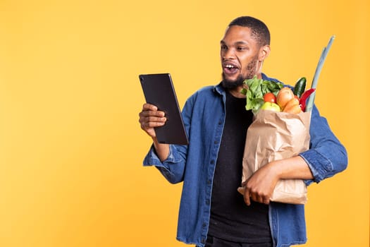 African american man checks his social media texts in studio, carrying a paper bag full of fruits and veggies from the local supermarket store. Confident relaxed guy having fun browsing the internet.