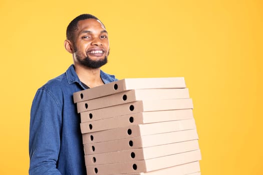 African american courier carrying huge stack of pizza boxes to deliver fast food order to customer, takeaway service in studio. Young cheerful deliveryman bringing takeout meal package.
