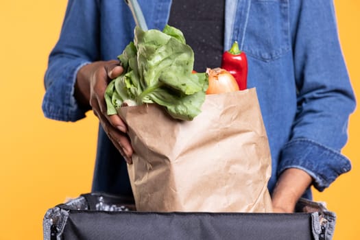 Male model courier emptying a paper bag full of fresh groceries, using backpack to bring local farmers market orders to clients. African american guy working in takeout industry. Close up.