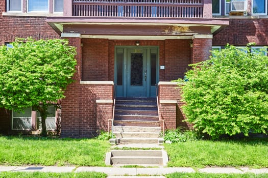 Aging red brick apartment in Fort Wayne, framed by lush foliage under a clear blue sky, embodies urban history and decay.